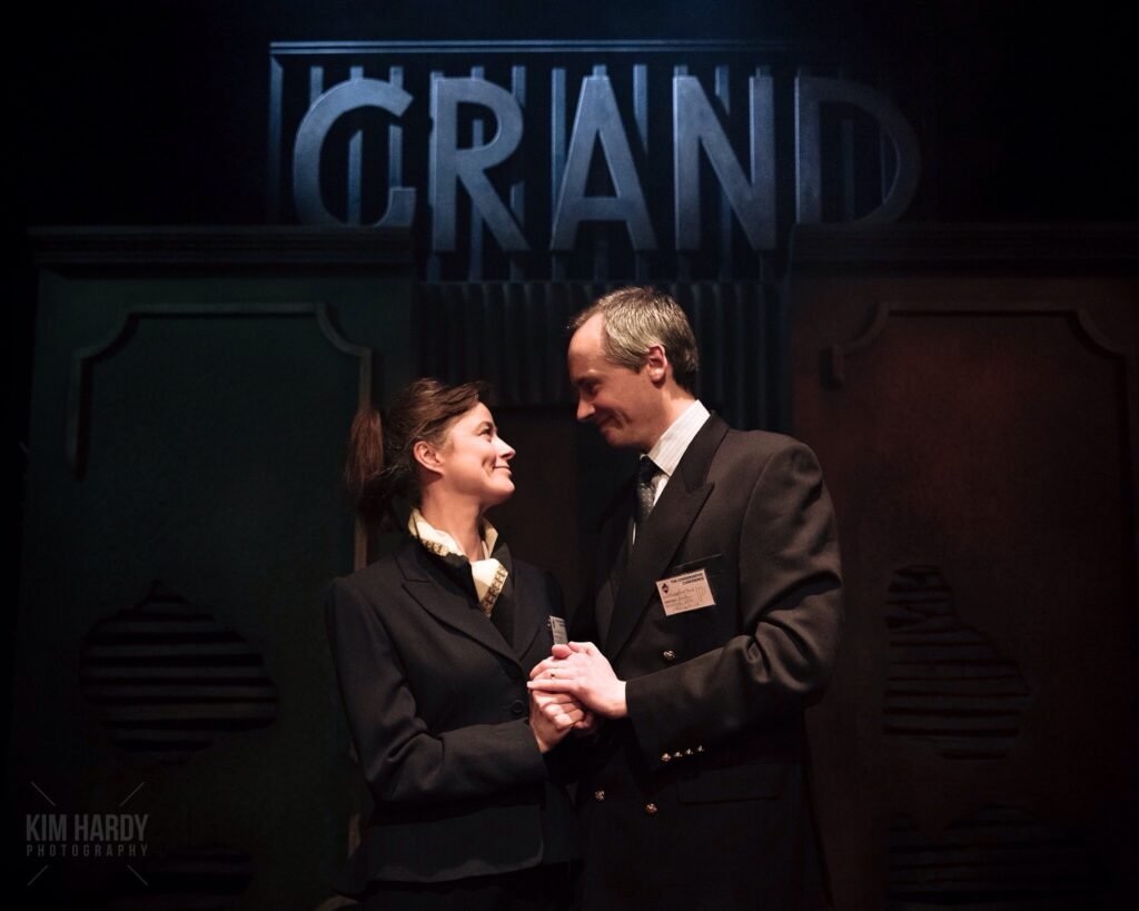 Two actors holding hands before a stage set reading GRAND, from The Bombing of the Grand Hotel