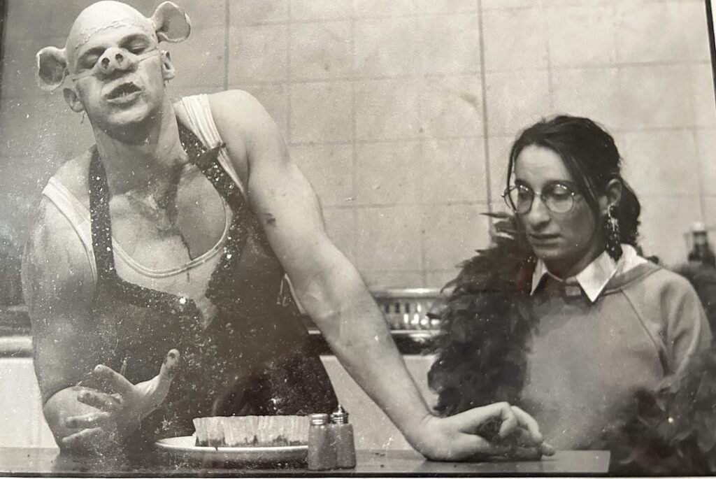 Production photo from Pig in a Poke — two actors at a kitchen counter, one wearing a pig mask, the other in glasses and a feather boa